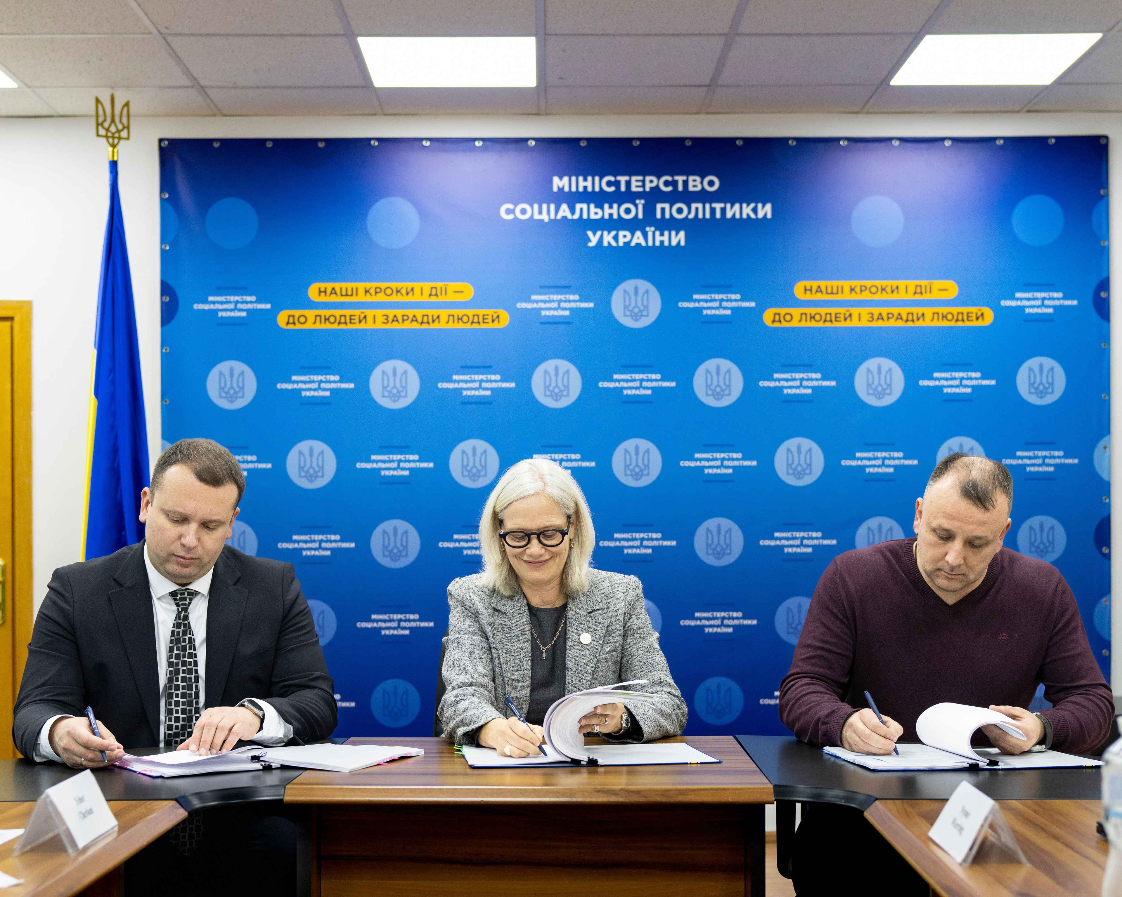 Дві особи, одна в білої сорочці, інша в темному костюмі, підписують документи на столі. Одна з них тримає ручку, а друга руку покладає на папери. На фоні видно комп'ютерну клавіатуру та годинник на руці одного з учасників.