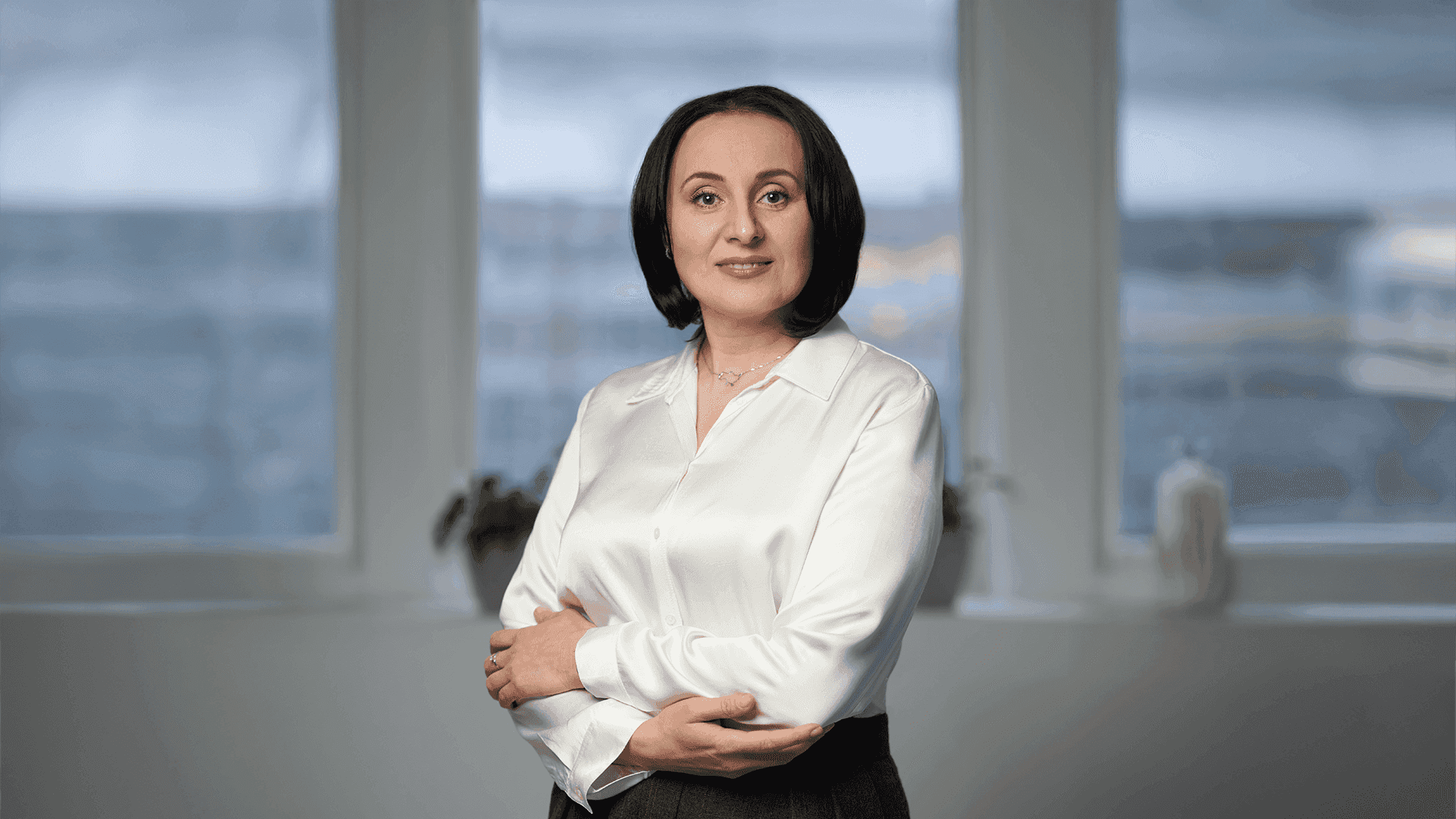 Жінка в білій блузці і сірих брюках, стоїть з схрещеними руками перед вікнами офісу. Це Жолнович Оксана Іванівна - Міністр соціальної політики України