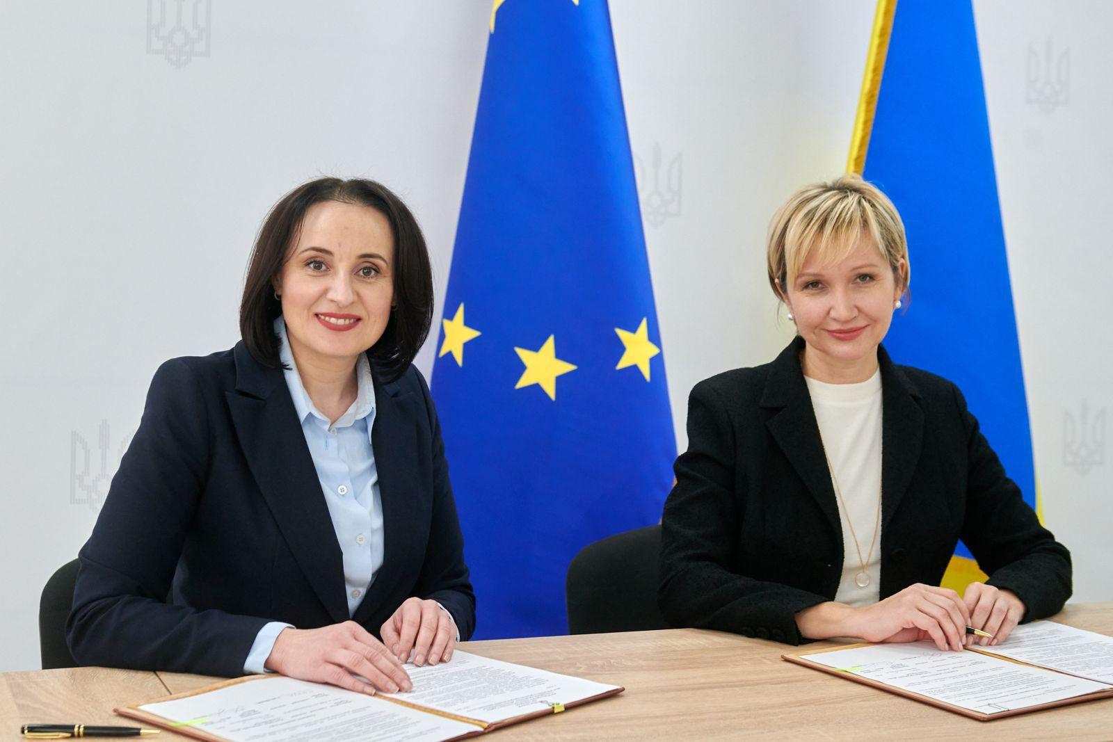 Жінки сидять за столом, підписуючи документи, на фоні двох прапорів — України та Європейського Союзу. Одна з жінок — Жолнович Оксана, інша — колега, обидві одягнені в костюми.