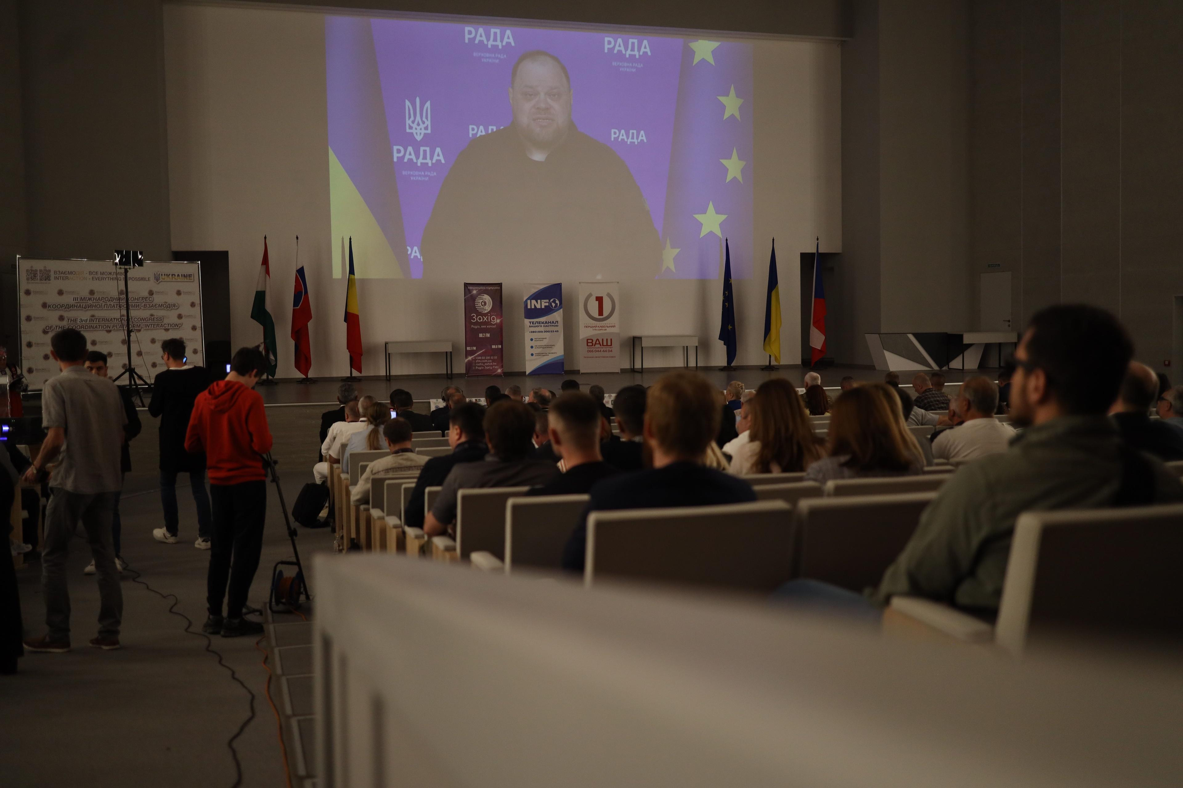 На зображенні багато людей сидять в залі. На екрані йде трансляція