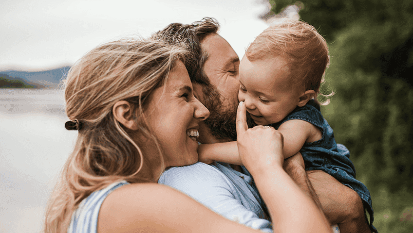 Сім'я на природі, батьки обіймають дитину, батько цілує її в щоку, а мати усміхається. Всі троє разом, виражаючи любов і прив’язаність. Сім'я на природі, батьки обіймають дитину, батько цілує її в щоку, а мати усміхається. Всі троє разом, виражаючи любов і прив’язаність.