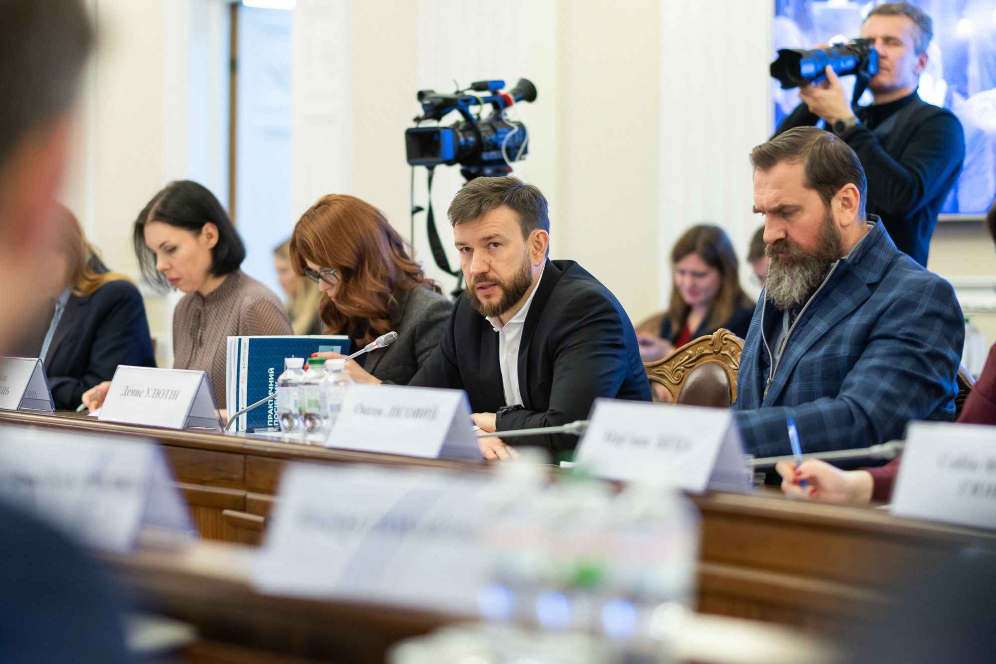 На фото чоловіки та жінки сидять під час наради На фото чоловіки та жінки сидять під час наради