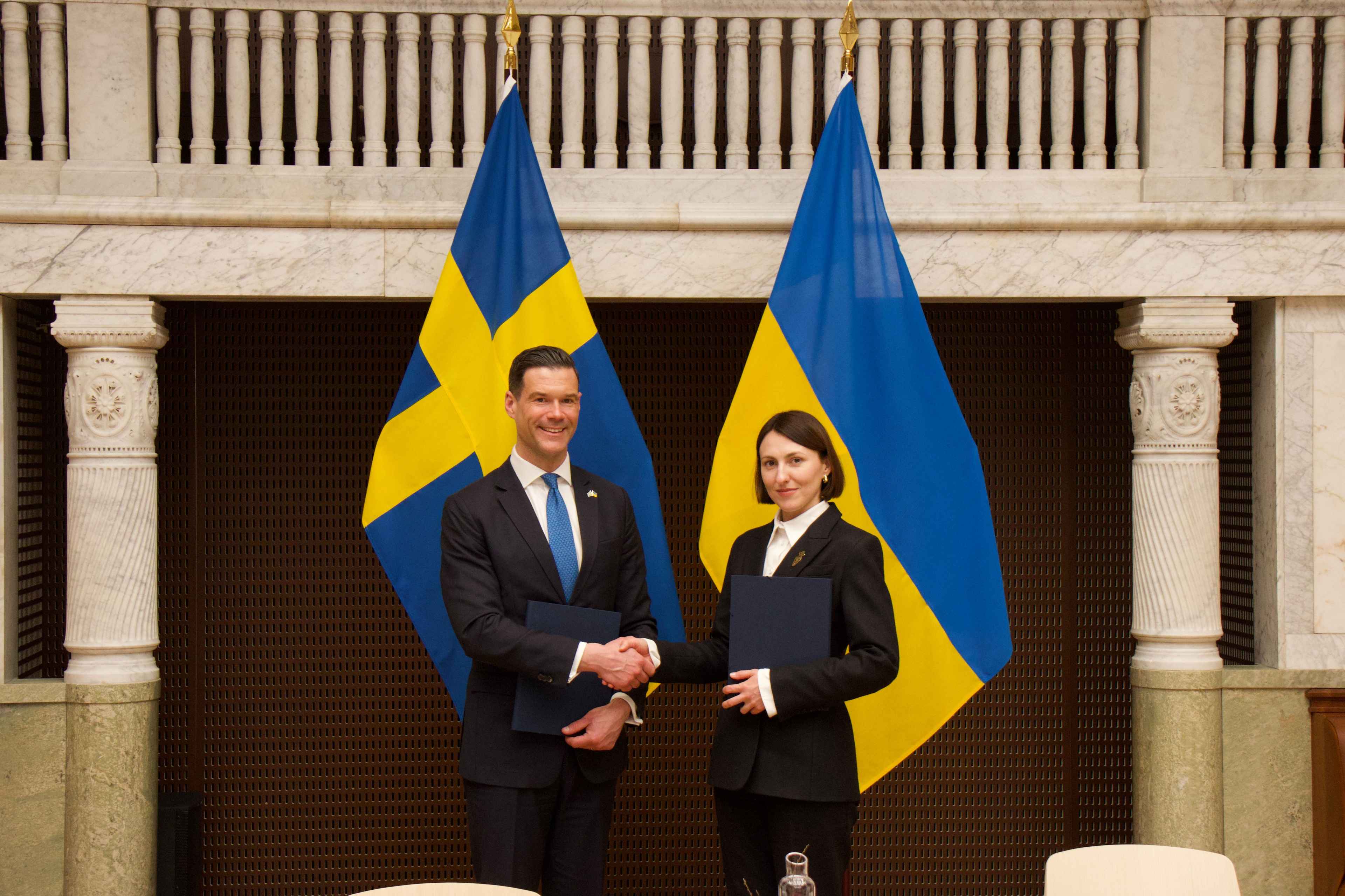 На фото чоловік та жінка вітаються за руку на фоні прапорів Швеції та України На фото чоловік та жінка вітаються за руку на фоні прапорів Швеції та України