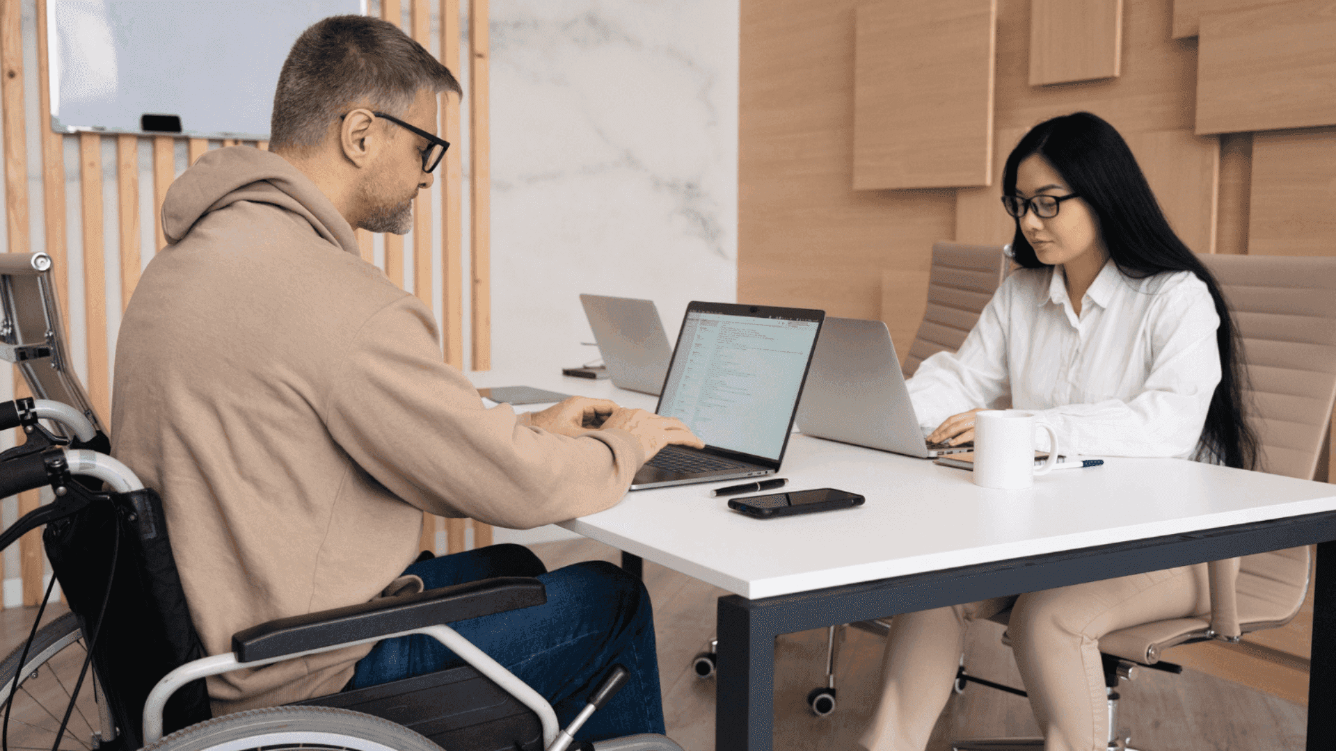 На фото чоловік з інвалідністю та жінка працюють за столом з ноутбками На фото чоловік з інвалідністю та жінка працюють за столом з ноутбками