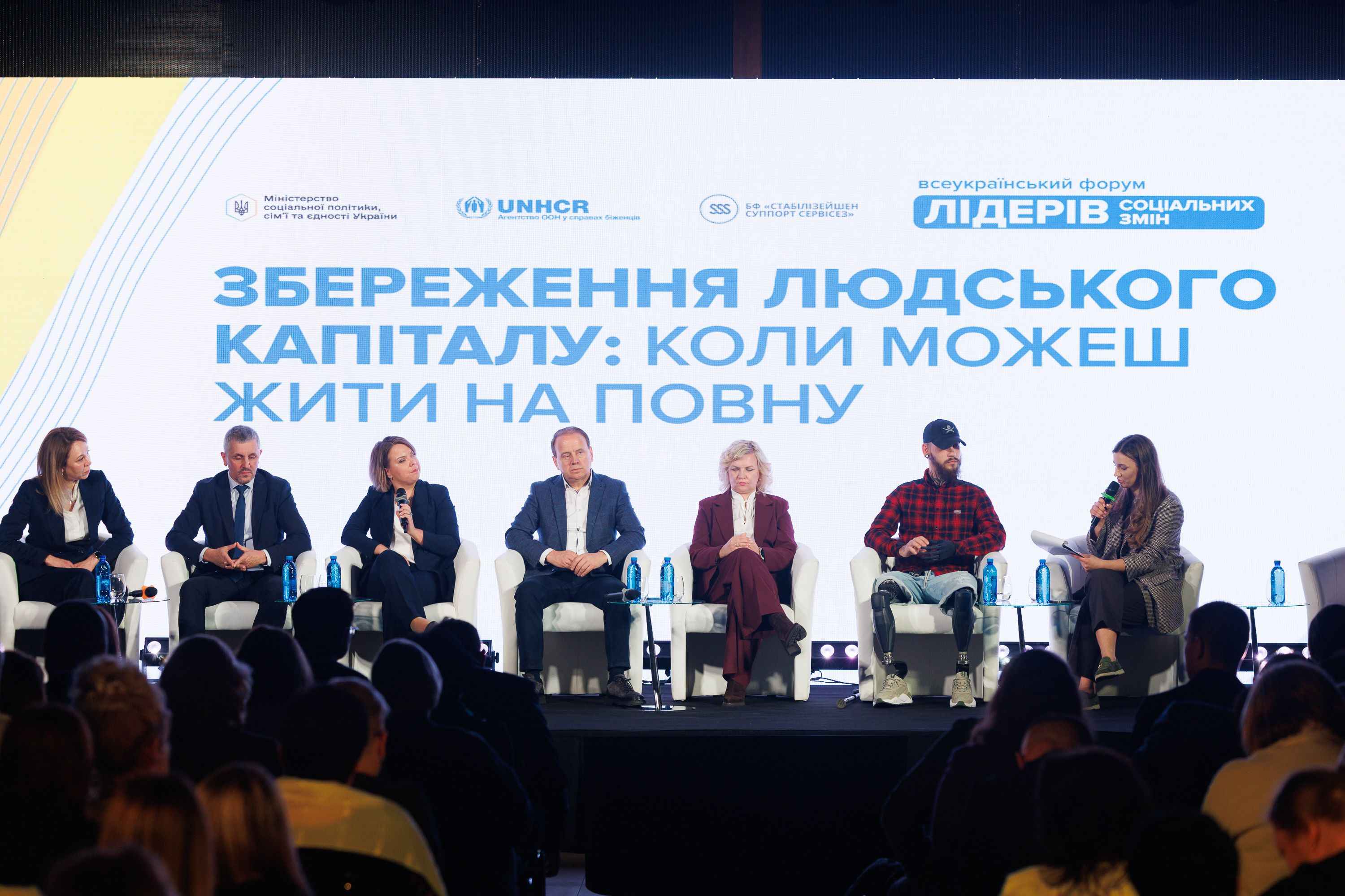 На фото жінки та чоловіки сидять на фоні сцени під час заходу На фото жінки та чоловіки сидять на фоні сцени під час заходу