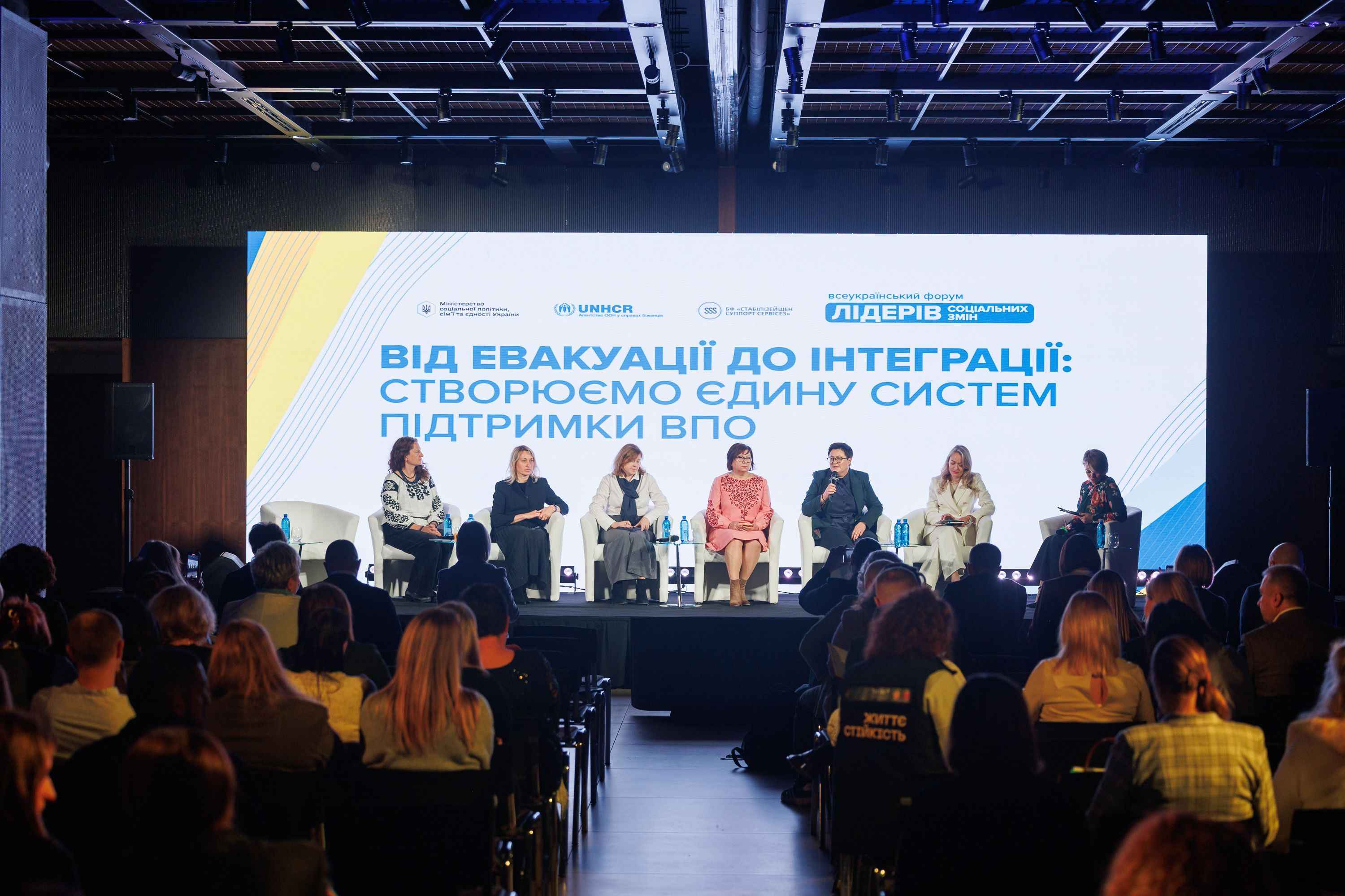 На фото жінки сидять на сцені на фоні банеру під час заходу На фото жінки сидять на сцені на фоні банеру під час заходу