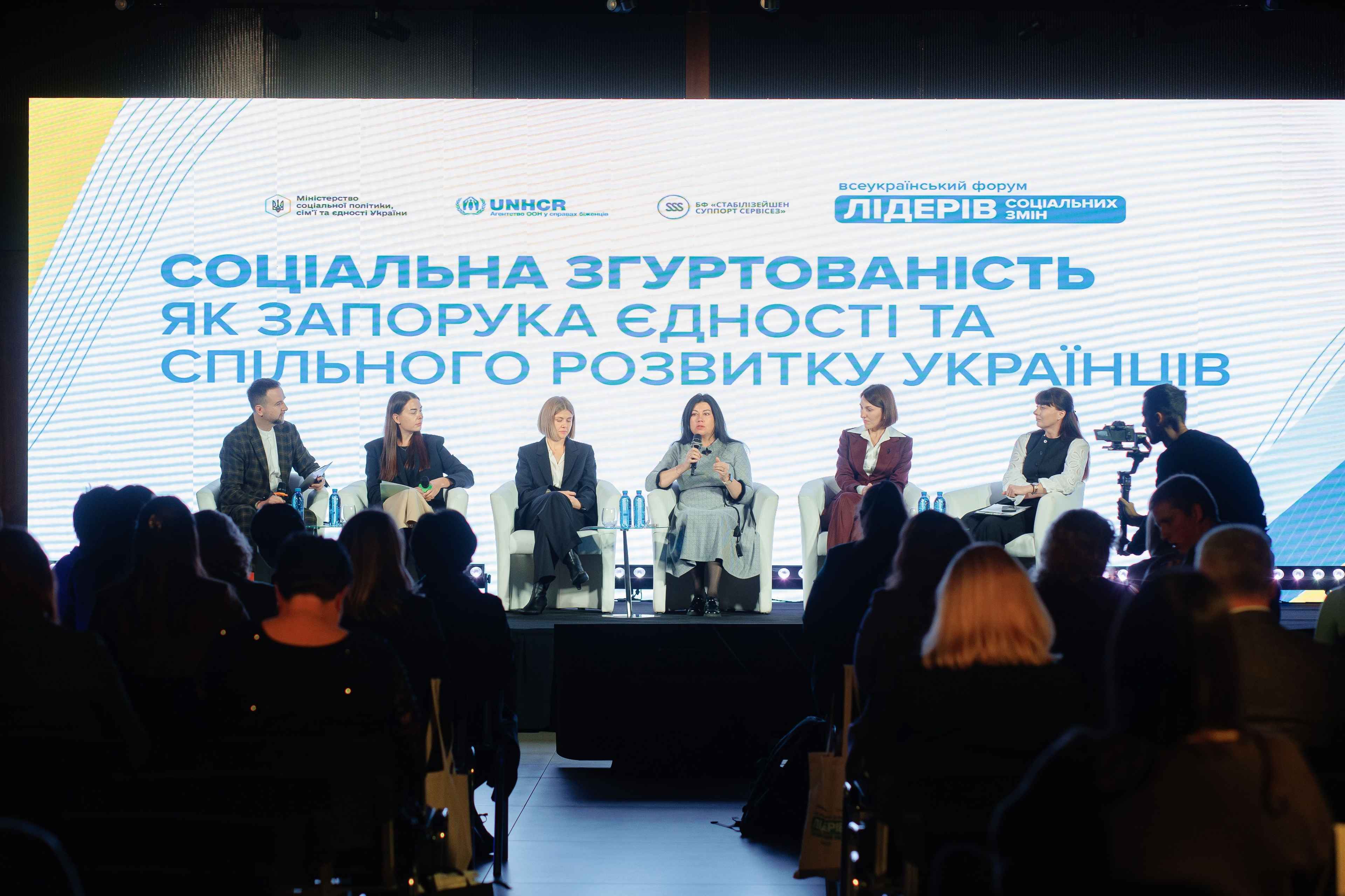 На фото чоловіки та жінки сидять на сцені під час заходу На фото чоловіки та жінки сидять на сцені під час заходу