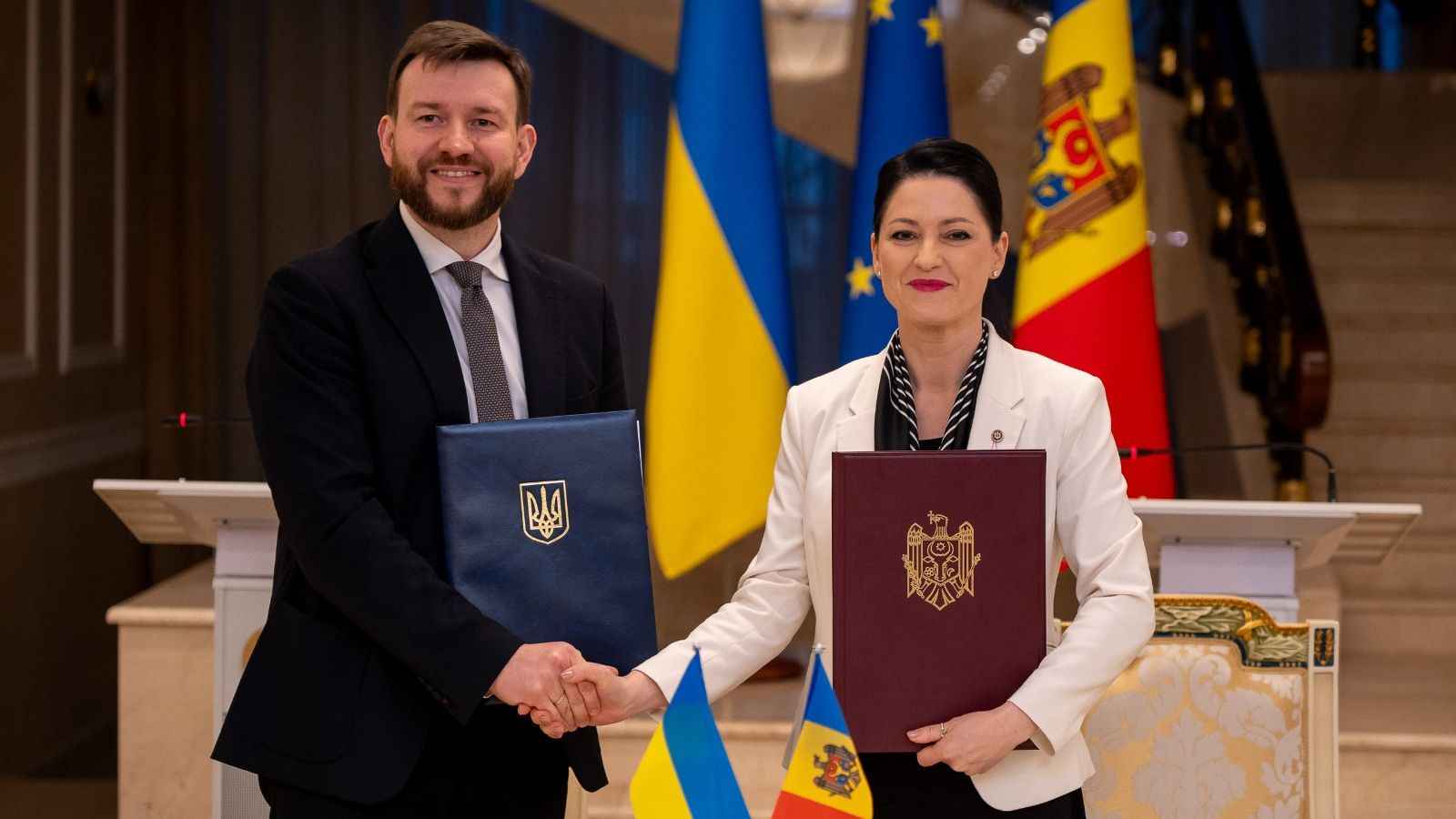 На фото чоловік та жінка вітаються за руку на фоні прапорів України та Республіки Молдова На фото чоловік та жінка вітаються за руку на фоні прапорів України та Республіки Молдова