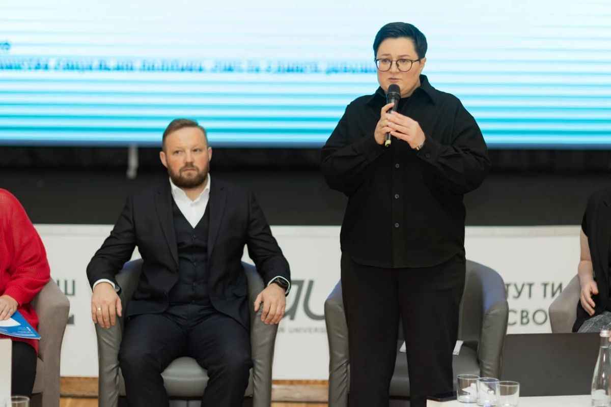 На фото чоловік та жінка у президії під час заходу, а жінка виступає з мікрофоном На фото чоловік та жінка у президії під час заходу, а жінка виступає з мікрофоном